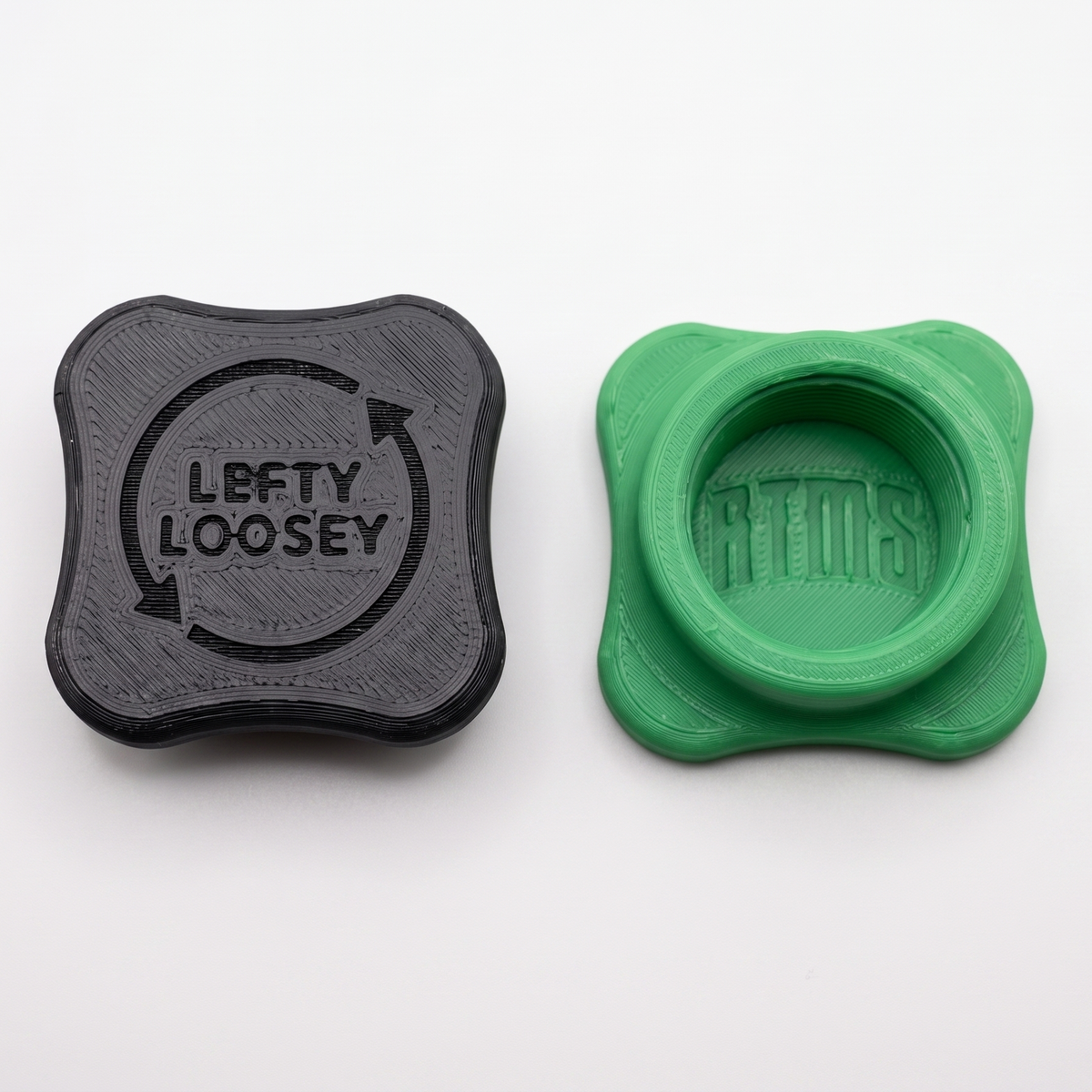 Two square-shaped 3D-printed plastic bottle openers with rounded corners on a white background. The left cap is dark black with the embossed text 'LEFTY LOOSEY' inside a circular arrow, and the right cap is bright green with the embossed text 'RTMS' inside a circular frame.